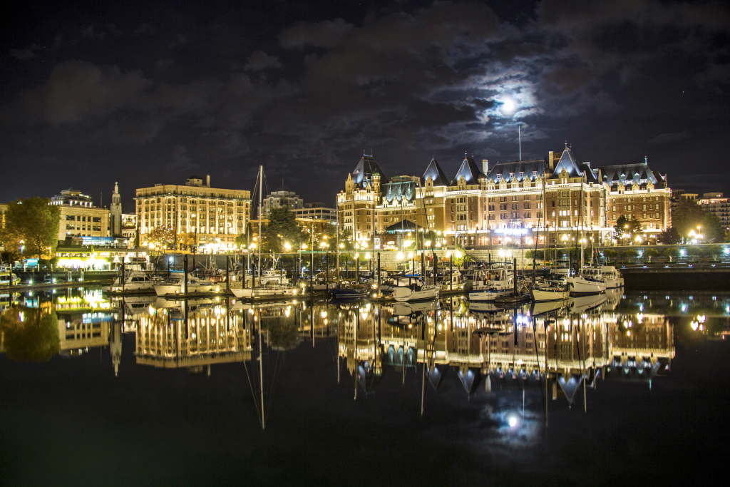 Victoria's inner harbour lit up at night. The parliament and musean are aglow and are mirrored horizontally alongside sail boats in the calm harbour water.
